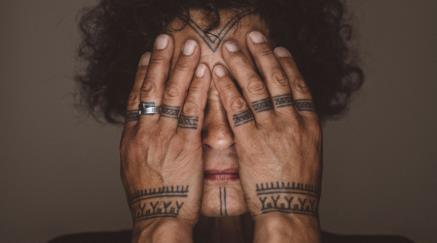 Person with tattooed hands covering eyes, showing rings and detailed ink.
