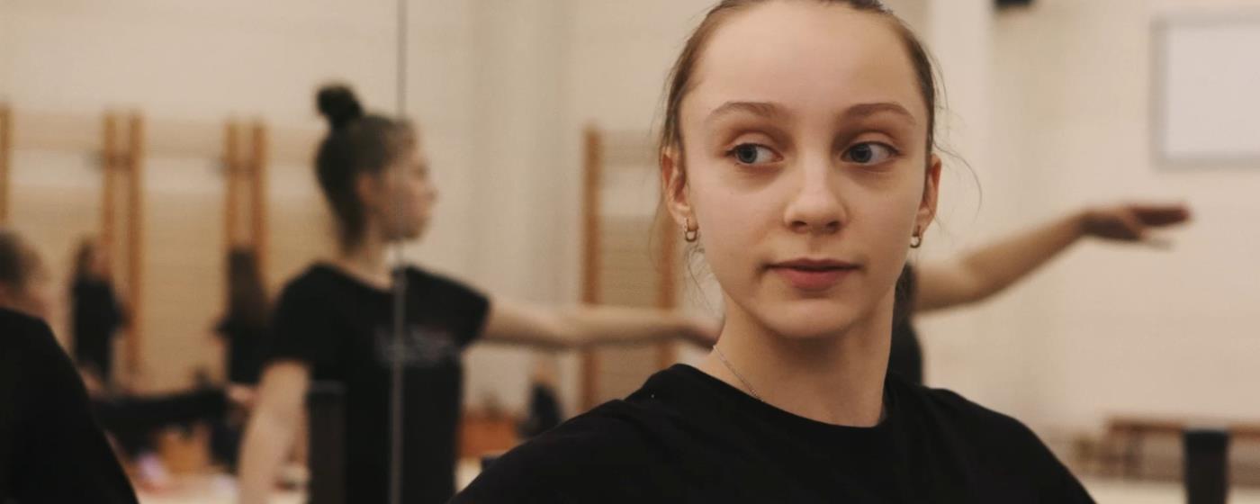 Young female dancer in studio with others practicing in background.