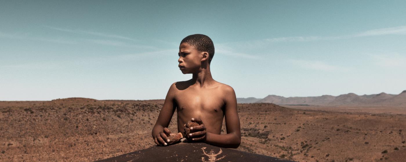Chlapec stojí za velkým kamenem a hledí do dáli přes pouštní krajinu.
