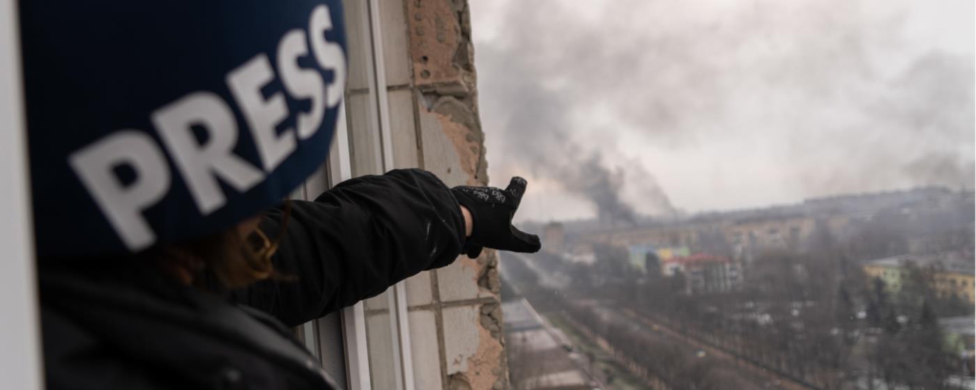 Journalist in a press vest/hat pointing at smoke rising over buildings.