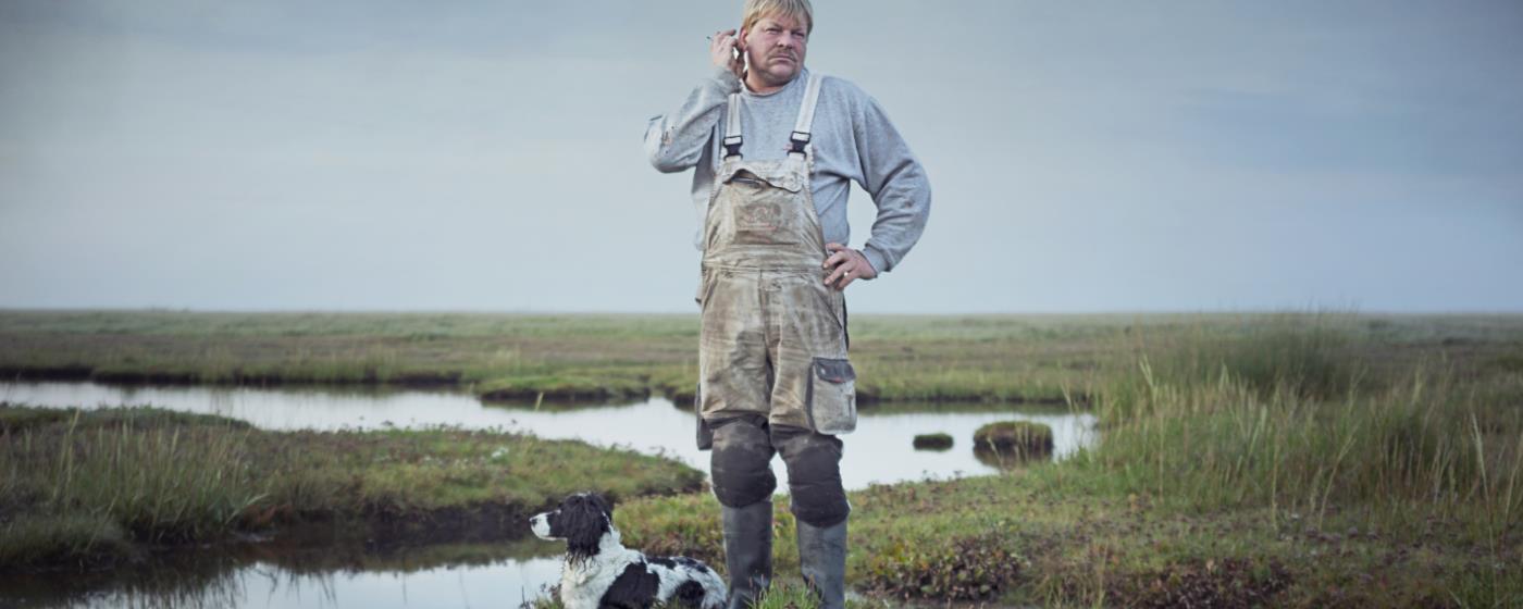 Man in overalls with dog standing by water in a field.