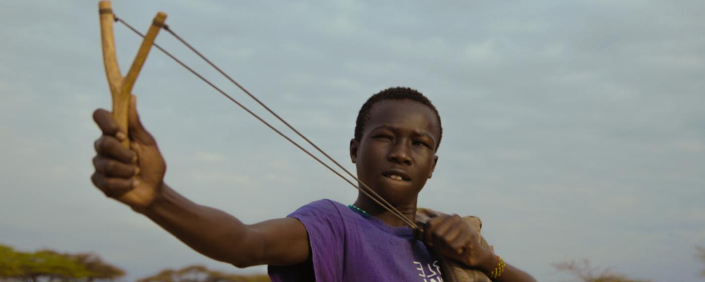 Boy aiming with a slingshot against a blue sky.
