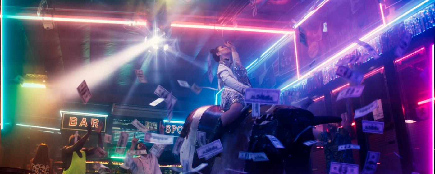 Barevně osvětlený interiér klubu s osobou sedící na mechanickém býkovi, kolem létají bankovky.