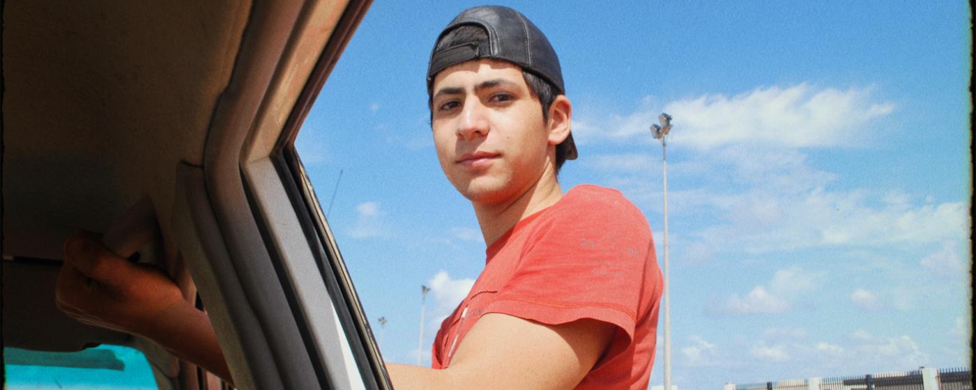 Young man in a red shirt and cap leaning on a car window looking at camera.