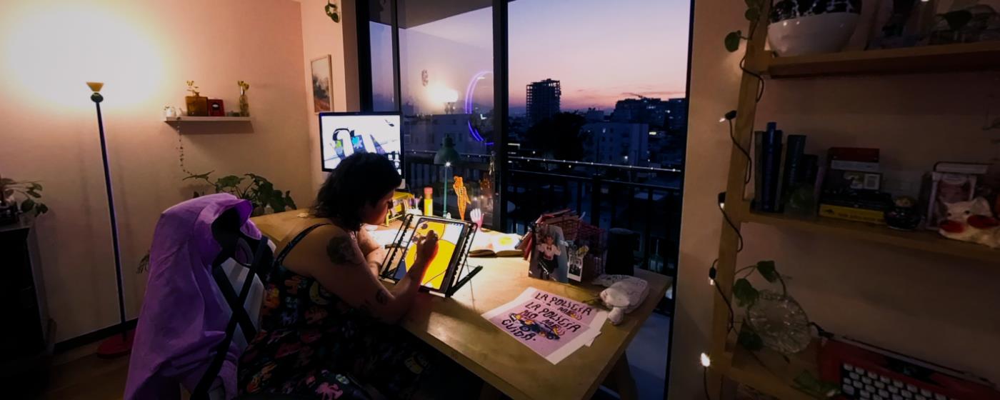 Osoba sedí u stolu a maluje v pokoji s výhledem na západ slunce ve městě.