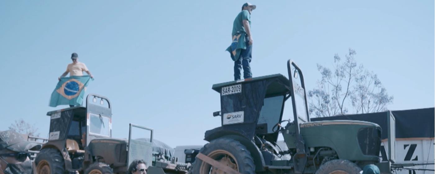 Dva muži stojí na střechách traktorů, jeden drží brazilskou vlajku.