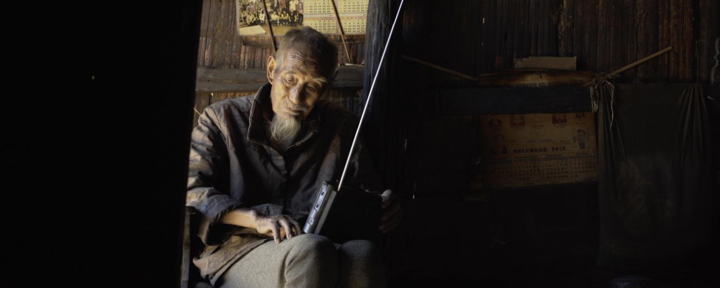 Elderly person holding a radio, seated inside a dim wooden cabin.