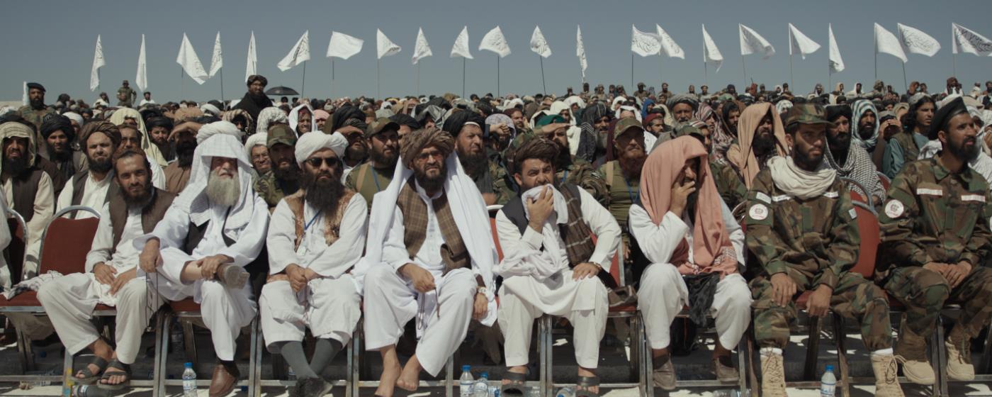Dav mužů v tradičním oblečení a vojáků sedí na tribuně pod řadou bílých vlajek.