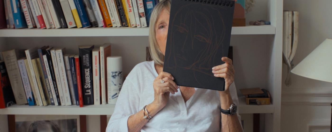 Starší žena drží skicák s kresbou obličeje, sedí před knihovnou.
