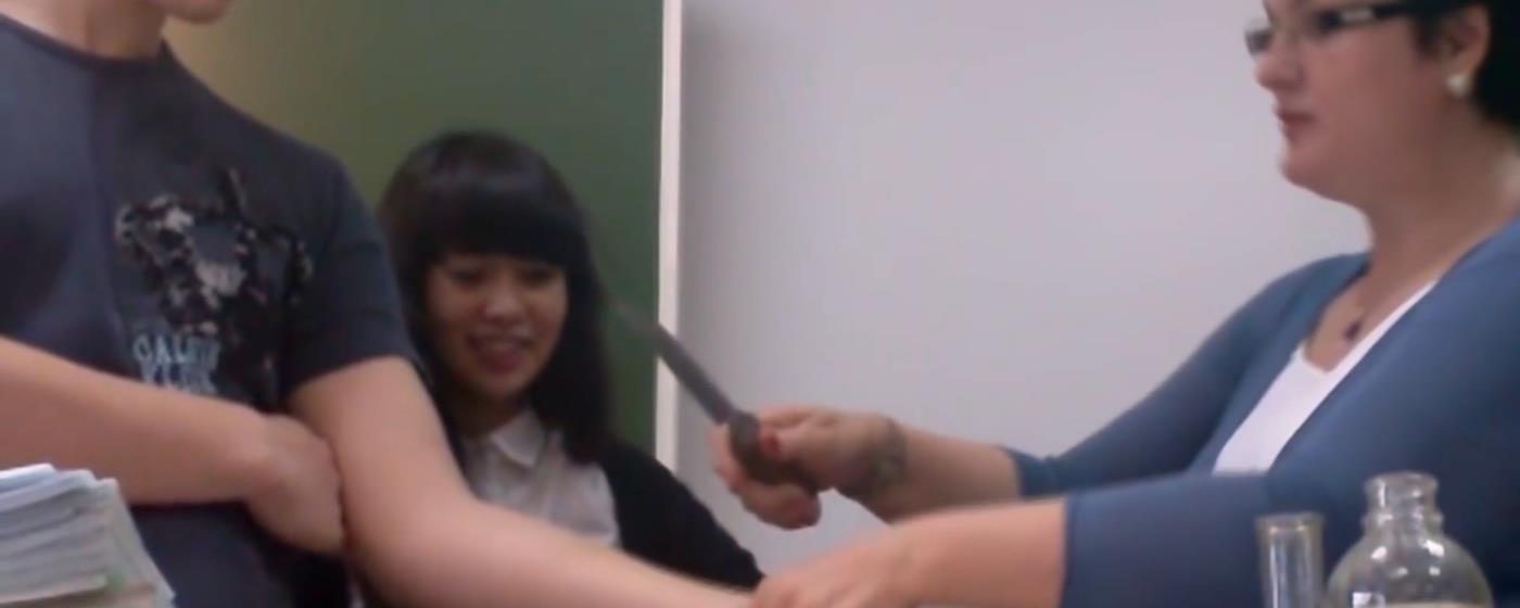 Teacher with knife close to a student's arm with one other student in background.
