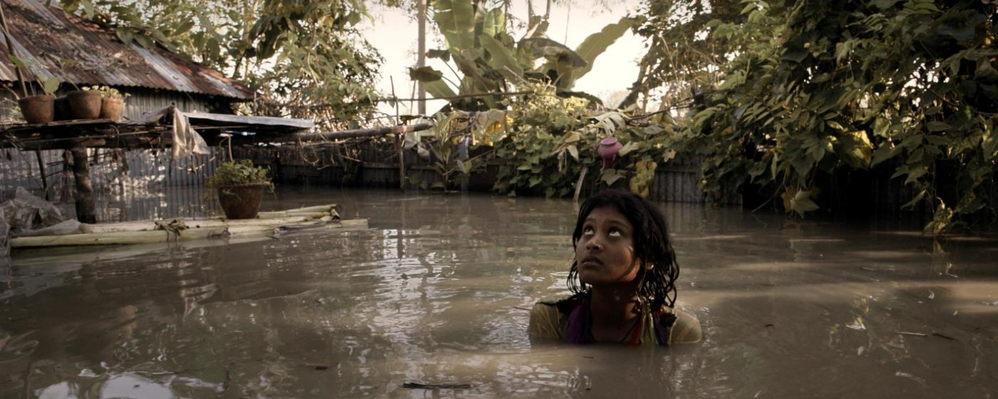 A girl looks up and wades through a flooded village with plants and a hut visible. 