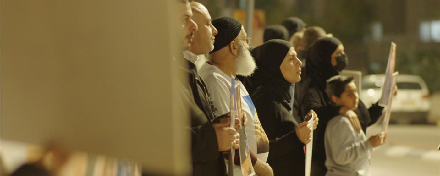 A diverse group of people standing in line, some wearing hijabs, holding posters. 