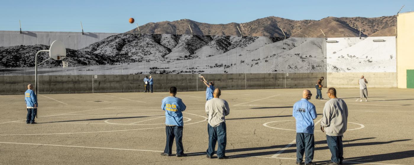 Vězni hrají basketbal na hřišti venku s horami v pozadí.
