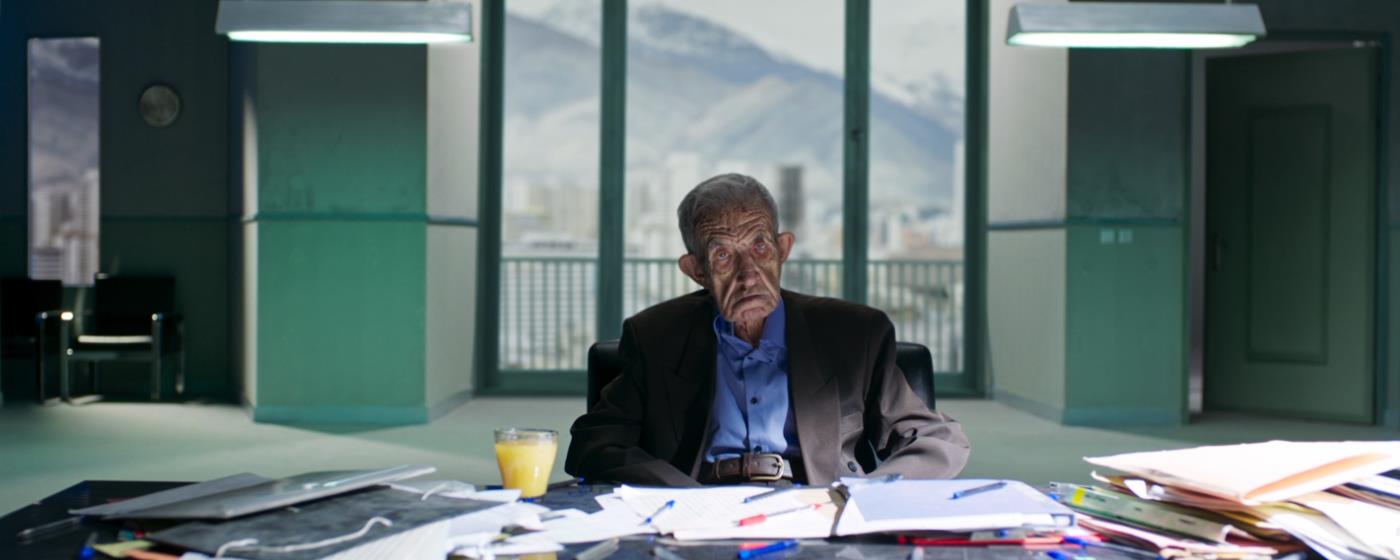 Starý muž sedí za kancelářským stolem, za oknem panorama hor.