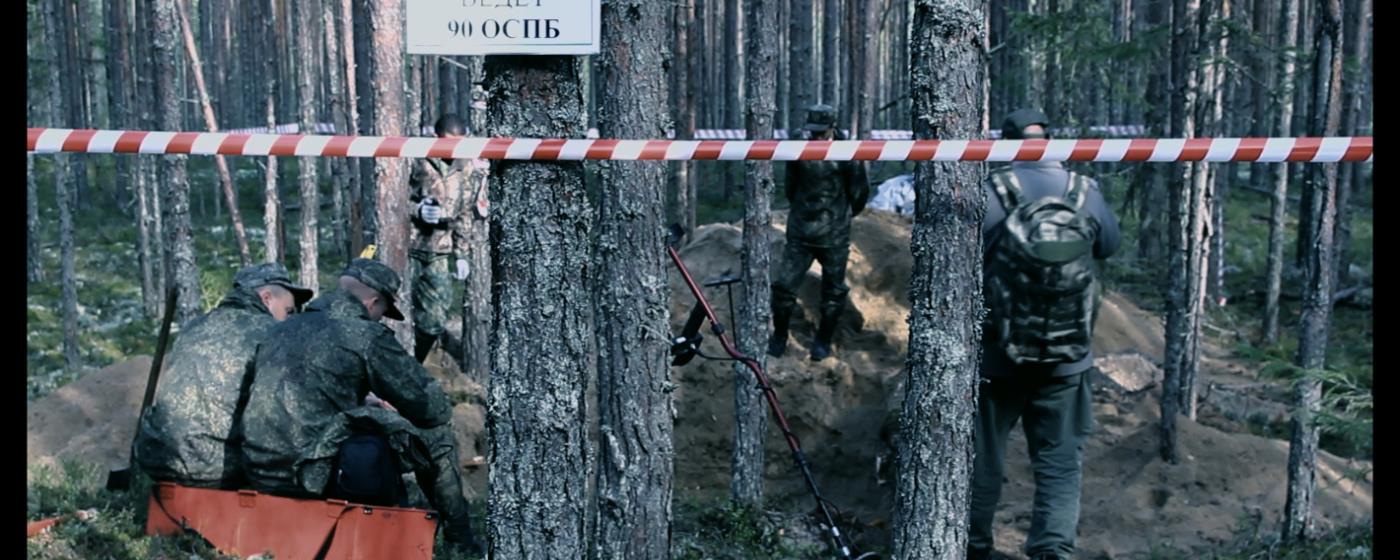 Skupina mužů v maskovacím oblečení v lese, prostor ohraničen výstražnou páskou.