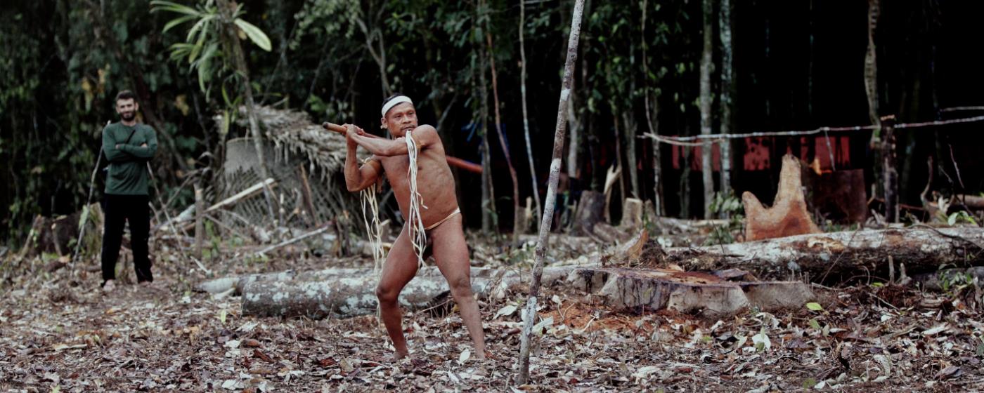 Muž v tradičním oděvu se rozmachuje s tyčí, druhý ho pozoruje ve vykáceném lese.