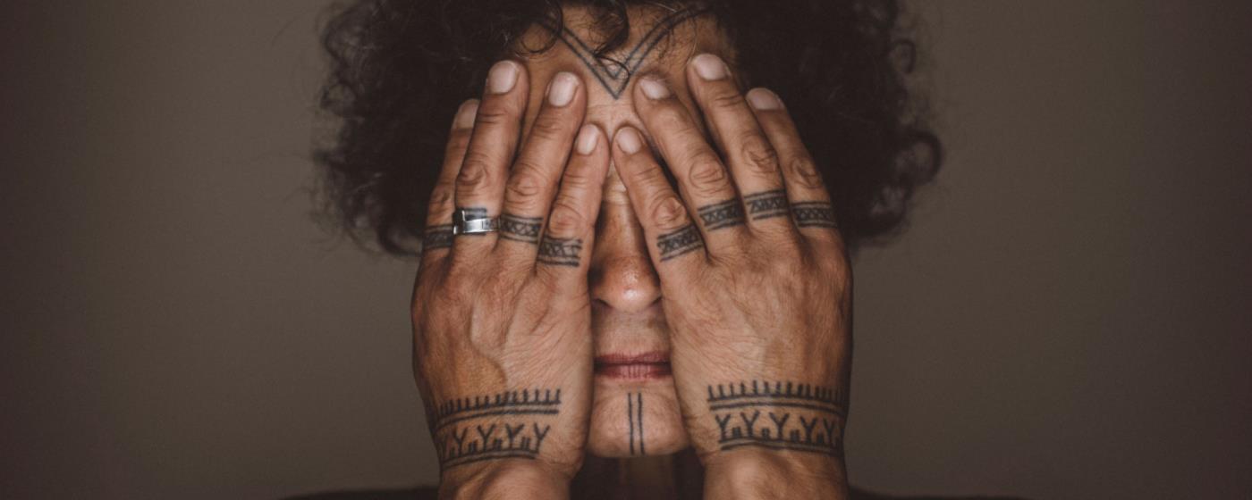 Person with tattooed hands covering eyes, showing rings and detailed ink.
