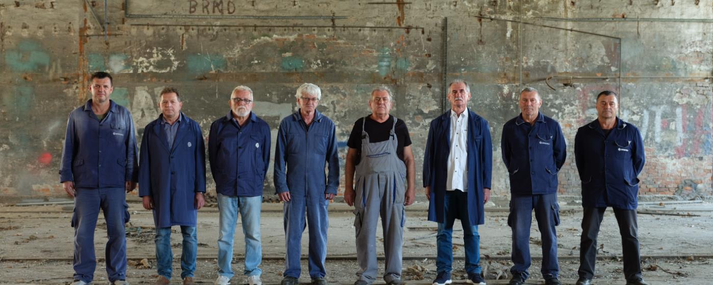 Eight men in blue uniforms are standing in a row in an abandoned building and are looking into the camera. 