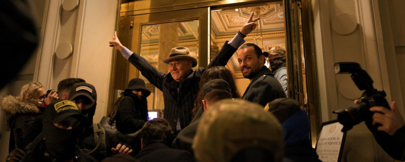 Man in a hat gesturing victoriously in a crowded doorway surrounded by cameras.