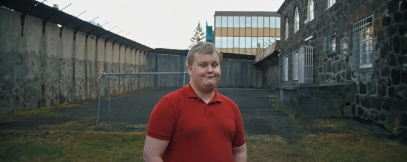 Muž v červeném tričku stojí před budovou s velkými okny za šera.