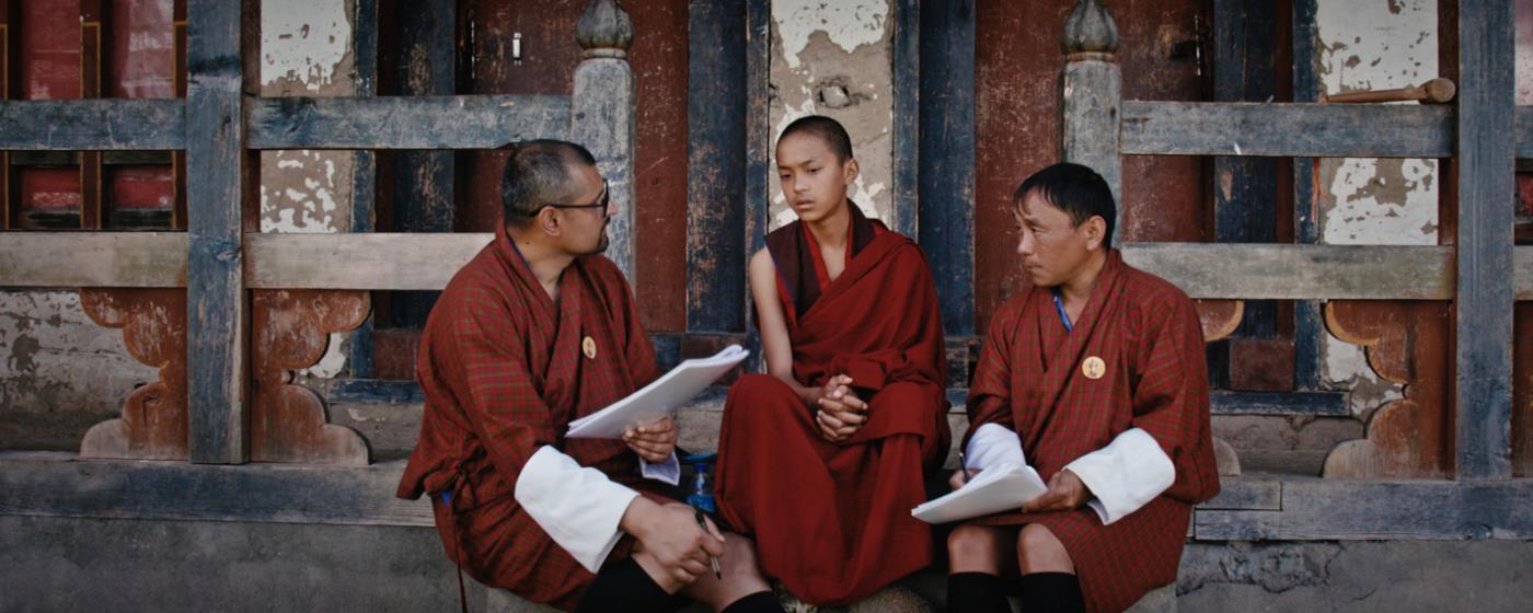 Two men and one boy in traditional robes sitting and talking by a building.
