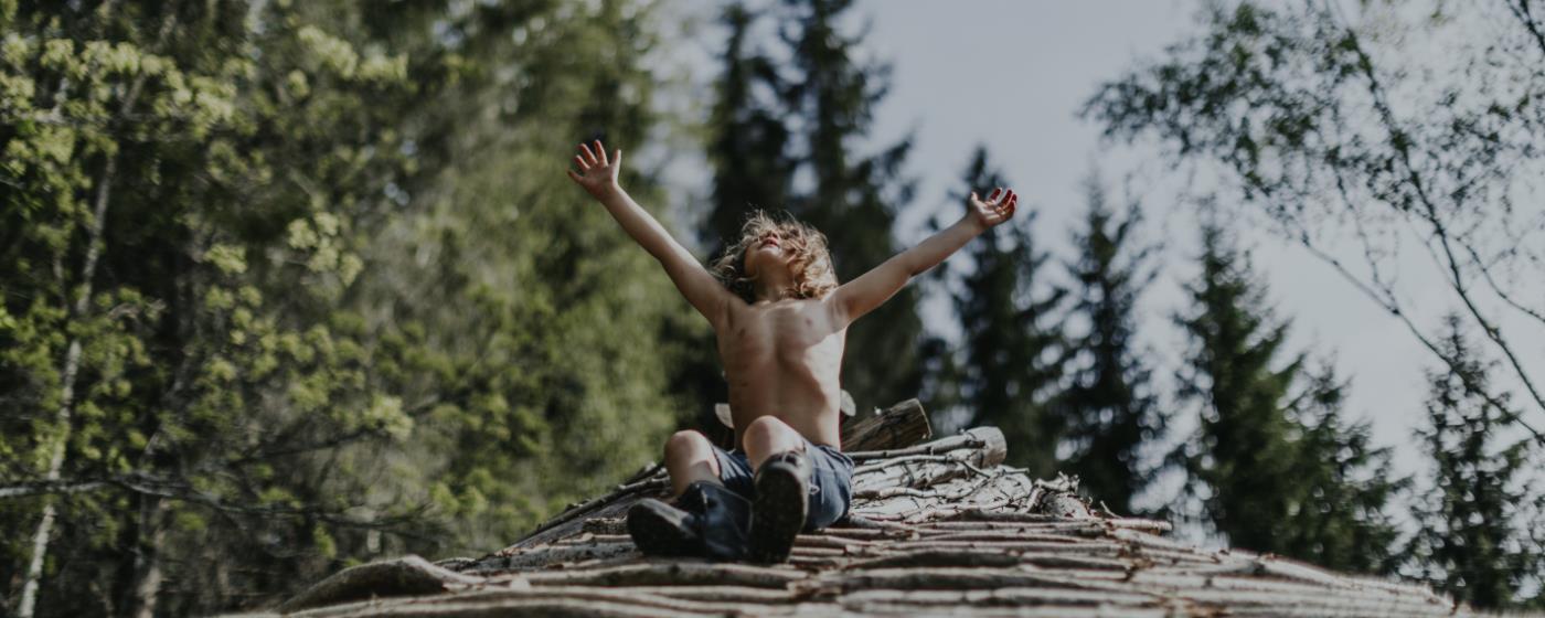 Chlapec s radostě zdviženýma rukama sedí na hromadě dříví v lese.