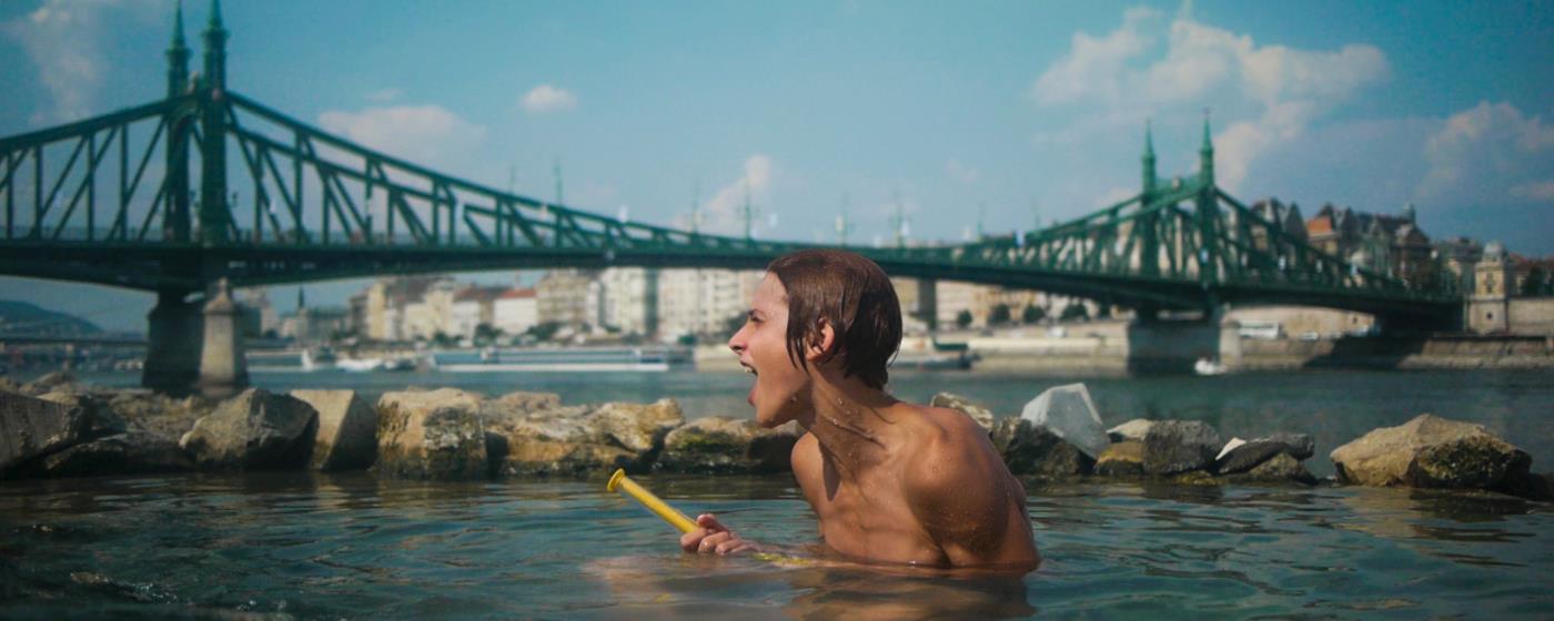 Chlapec s radostným výrazem se koupe v řece, v pozadí most a silueta města.