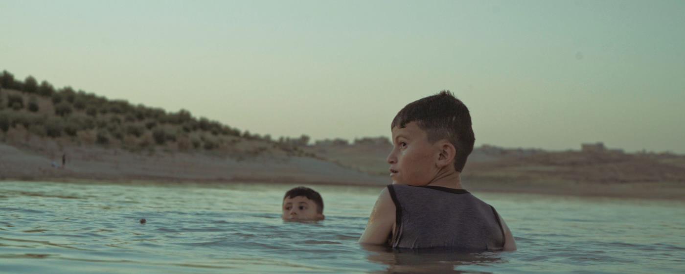 Dva chlapci plavou ve vodě, jeden z nich se dívá přes rameno.