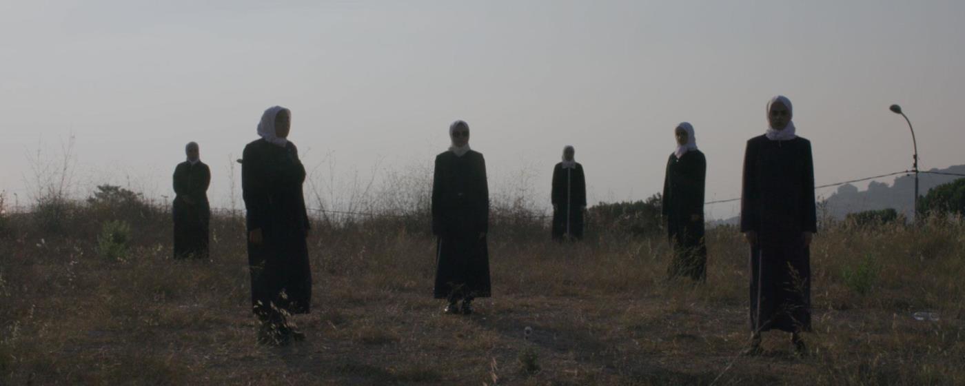 Several figures in dark robes, white headresses standing in a barren field.