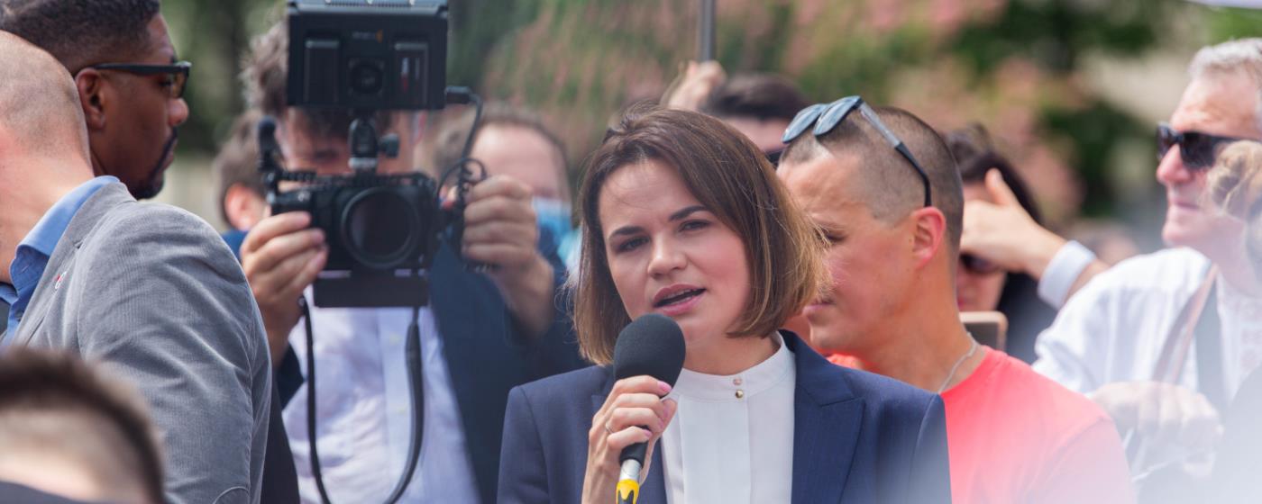 Woman speaking into a microphone at an outdoor event with a camera nearby.