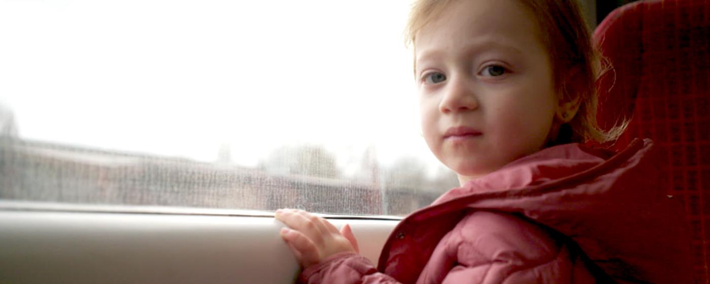 Young child in a pink jacket in car turning from window at camera.