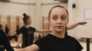 Young female dancer in studio with others practicing in background.