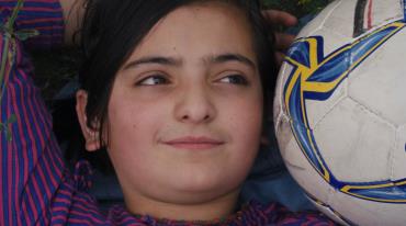 Smiling girl lying on grass with a soccer ball near her head.
