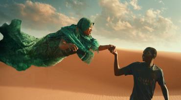Woman in green floating above desert, holding hands with a guiding, smiling boy.