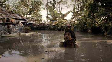 A girl looks up and wades through a flooded village with plants and a hut visible. 