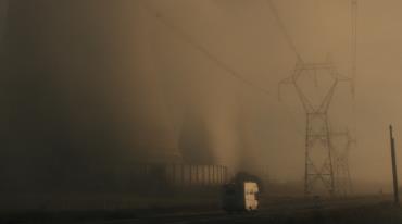 Elektrárna v mlze, siluety chladících věží a elektrické stožáry, vpředu projíždí nákladní vůz.