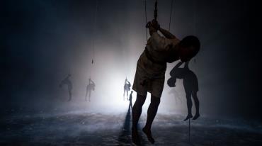Silhouettes of figures hanging by hands behind back in a misty room with ethereal lighting. 