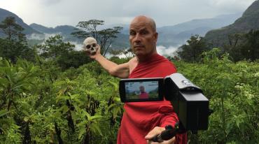 Muž drží lebku a natáčí se kamerou v tropické krajině.