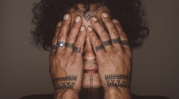 Osoba s tetováním na prstech si zakrývá obličej rukama.