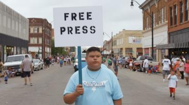 Chlapec drží ceduli s nápisem "FREE PRESS" na ulici plné lidí.
