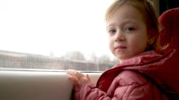 Malá dívka v růžové bundě sedí u okna vlaku, venku je zamračeno.