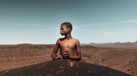 Chlapec stojí za velkým kamenem a hledí do dáli přes pouštní krajinu.
