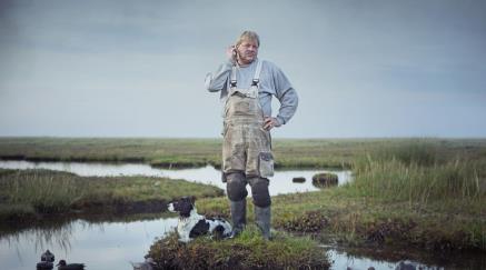 Man in overalls with dog standing by water in a field.