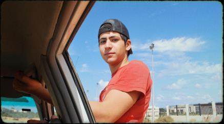 Young man in a red shirt and cap leaning on a car window looking at camera.