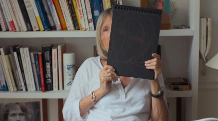 Starší žena drží skicák s kresbou obličeje, sedí před knihovnou.