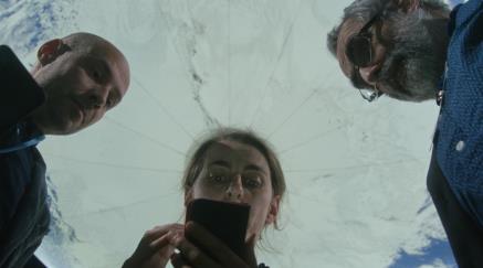 Tři lidé shlížejí dolů na obrazovku telefonu, nad nimi maketa planety Země.