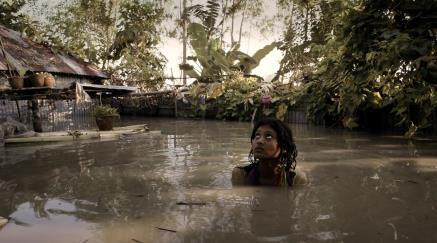 Dívka stojící ve vodě před zaplaveným domem s vegetací.