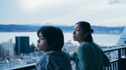 Woman and teen child looking at a cityscape from a high balcony in overcast setting.