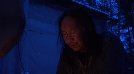 Man with long hair squinting, night setting with blue lighting, flame light against face. 