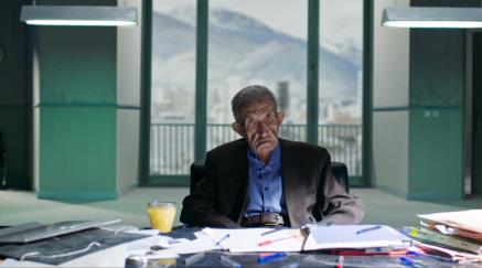 Starý muž sedí za kancelářským stolem, za oknem panorama hor.